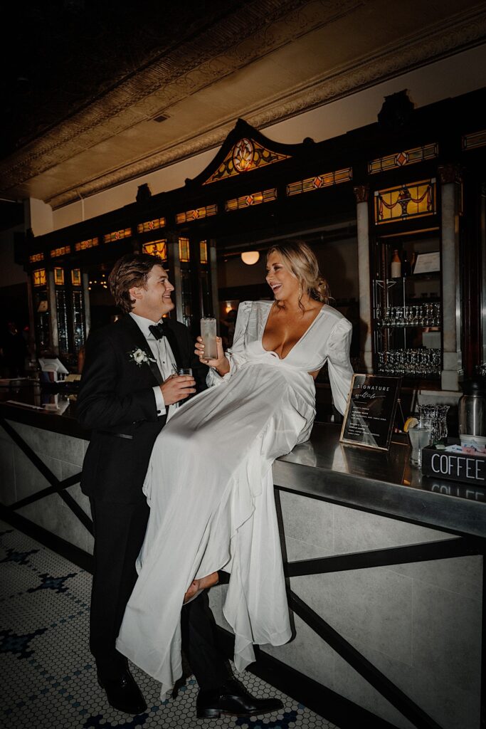 A bride sits on a bar as the groom stands next to her, they are both holding drinks and they are smiling at one another
