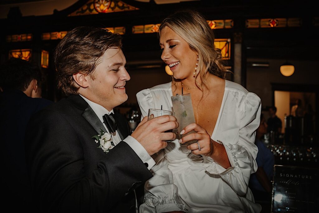 A bride and groom clink their glasses together while smiling at one another at the bar during their indoor wedding reception at Venue CU in Champaign Illinois