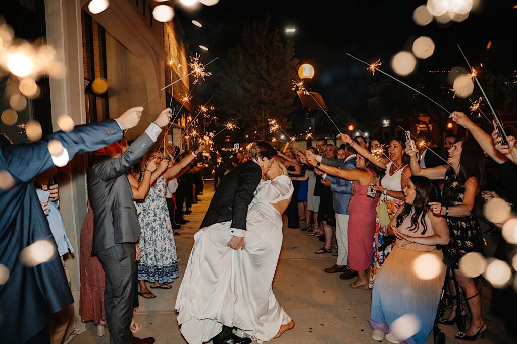 A bride is kissed and dipped by the groom while their guests around them form a tunnel with sparklers as they exit their wedding at Venue CU in Champaign Illinois