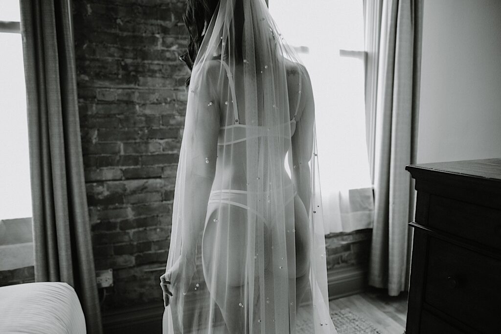 Black and white photo of a woman in lingerie and a veil standing and facing away from the camera while holding her veil, in front of her is a brick wall and two windows, photo taken during a bridal boudoir session