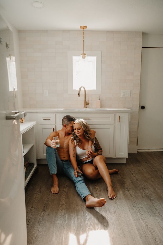 A man and woman sit on the floor in the kitchen and embrace while drinking water
