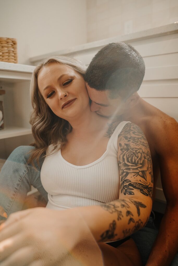 A shirtless man kisses a woman on the neck from behind while they sit on the floor in a kitchen