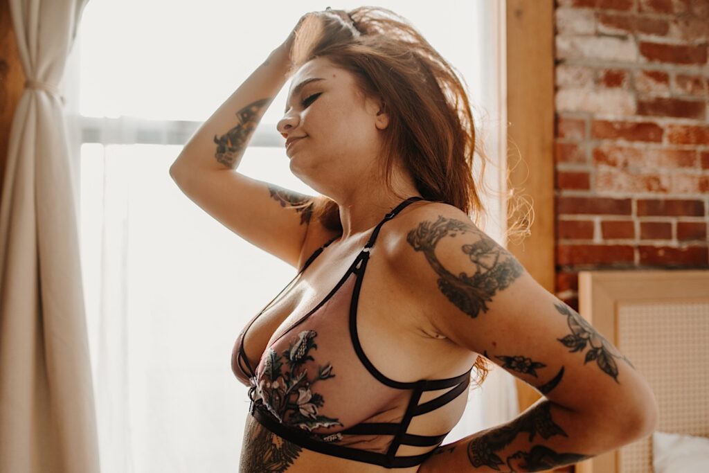 A tattooed woman in lingerie stands in front of a window and touches her hair with one of her hands while having boudoir photos taken of her in Chicago