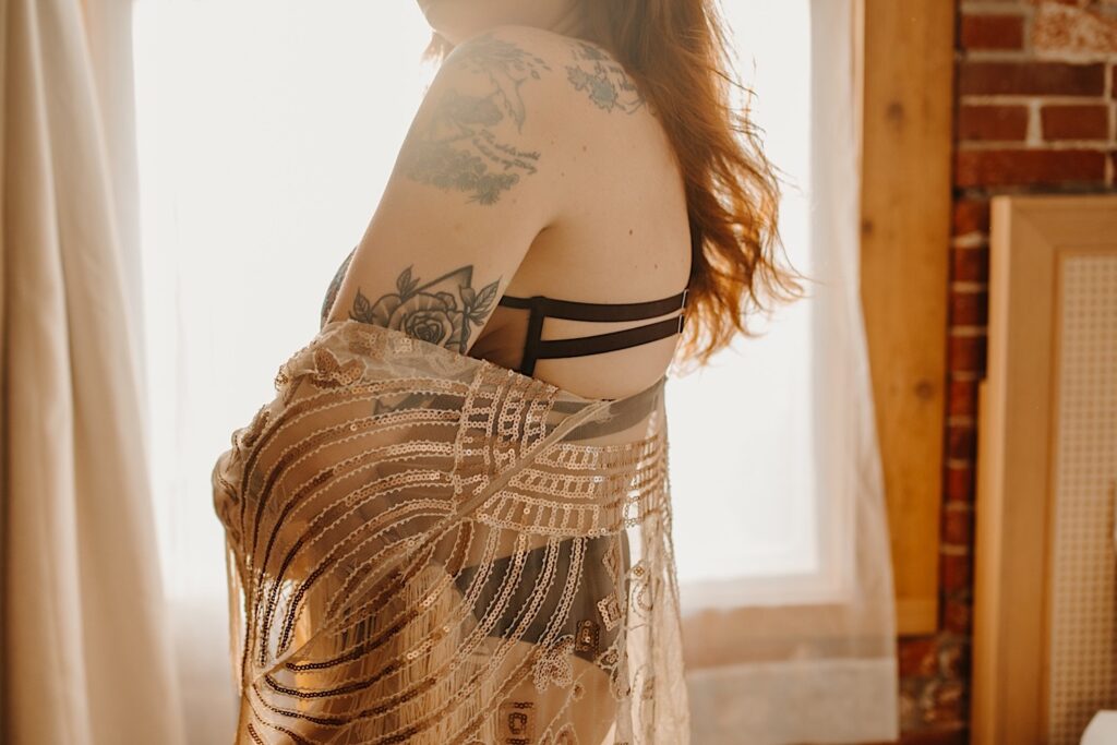 A tattooed woman in lingerie stands in front of a window and drapes a piece of fabric in front of her while having boudoir photos taken of her in Chicago