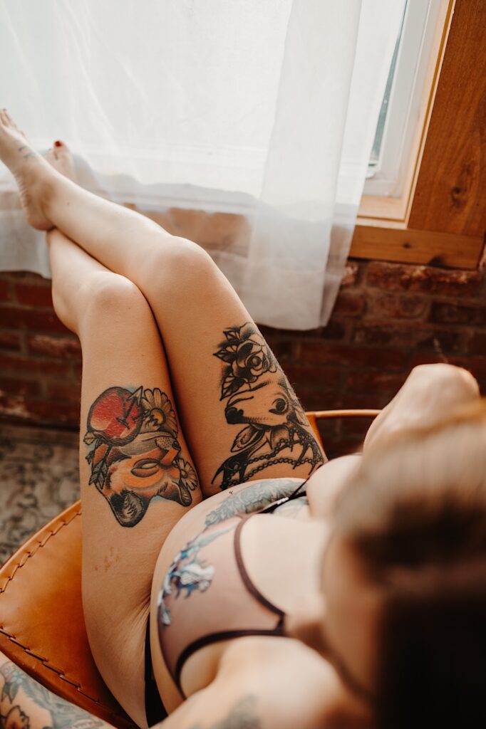A tattooed woman sits in a chair in front of a window while wearing lingerie and resting her feet on the windowsill