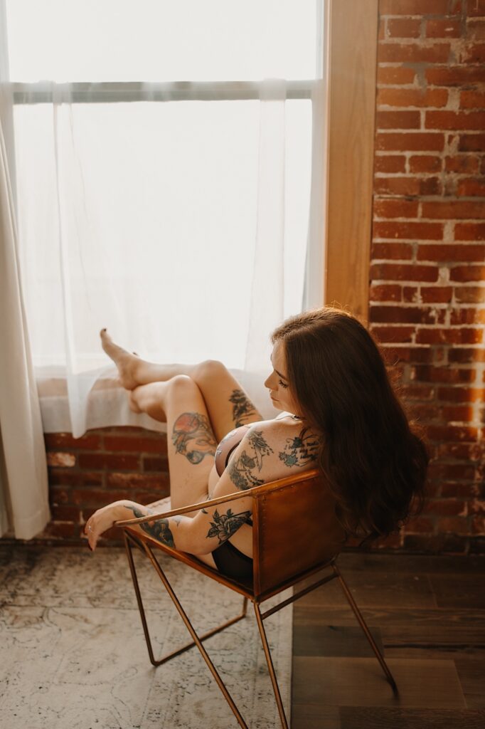 A tattooed woman sitting in a chair faces away from the camera and looks over her shoulder while resting her feet on the windowsill in front of her