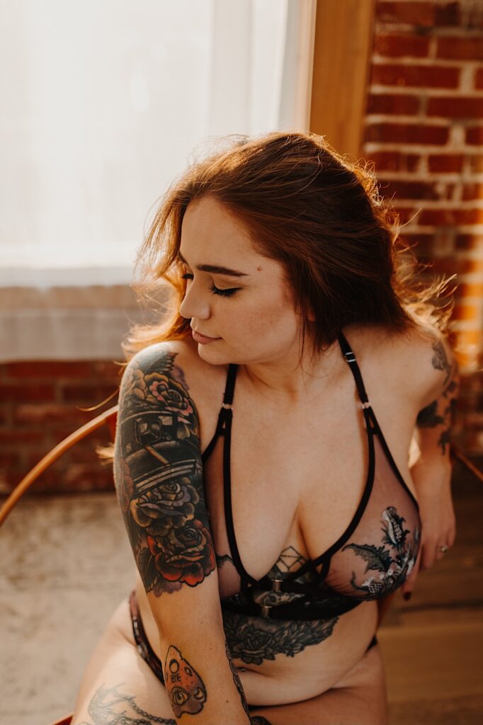 A tattooed woman in lingerie sits and faces the camera while looking to her left towards a window behind her