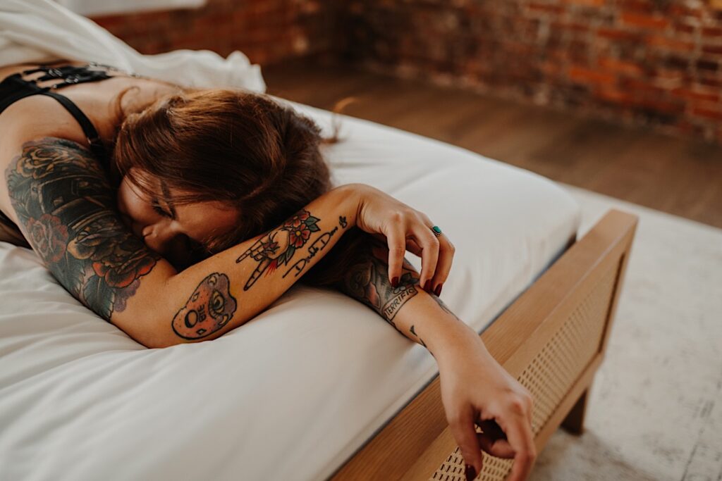 A tattooed woman lays on her stomach wearing lingerie and touches her arm with her eyes closed while having boudoir photos taken of her in Chicago