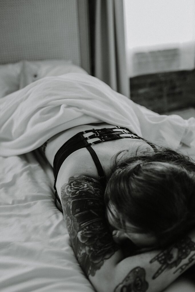 Black and white photo of a tattooed woman laying facedown on a bed with a sheet covering the bottom half of her body