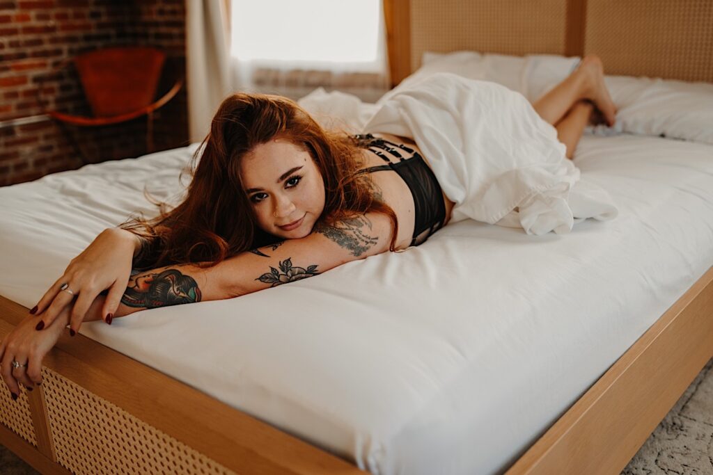 A tattooed woman lays on her stomach on a bed while wearing lingerie as a sheet covers the bottom half of her body, she is looking up at the camera while having boudoir photos taken of her in Chicago