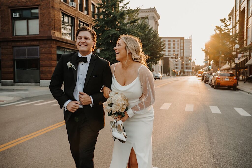 A bride and groom smile walking with their arms locked down a street in Champaign Illinois at sunset outside of their wedding venue, Venue CU
