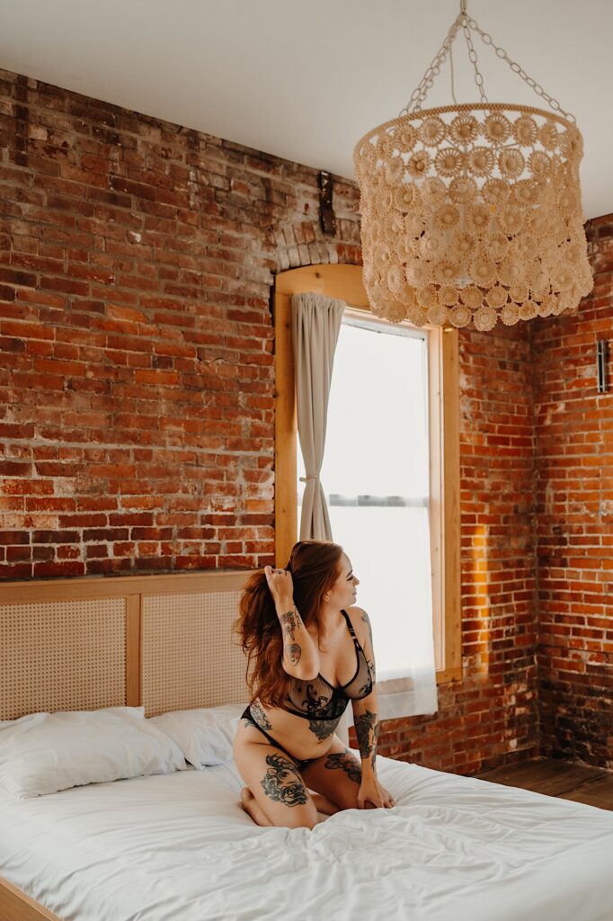 A woman kneels on a bed in a brick room while wearing lingerie and playing with her hair