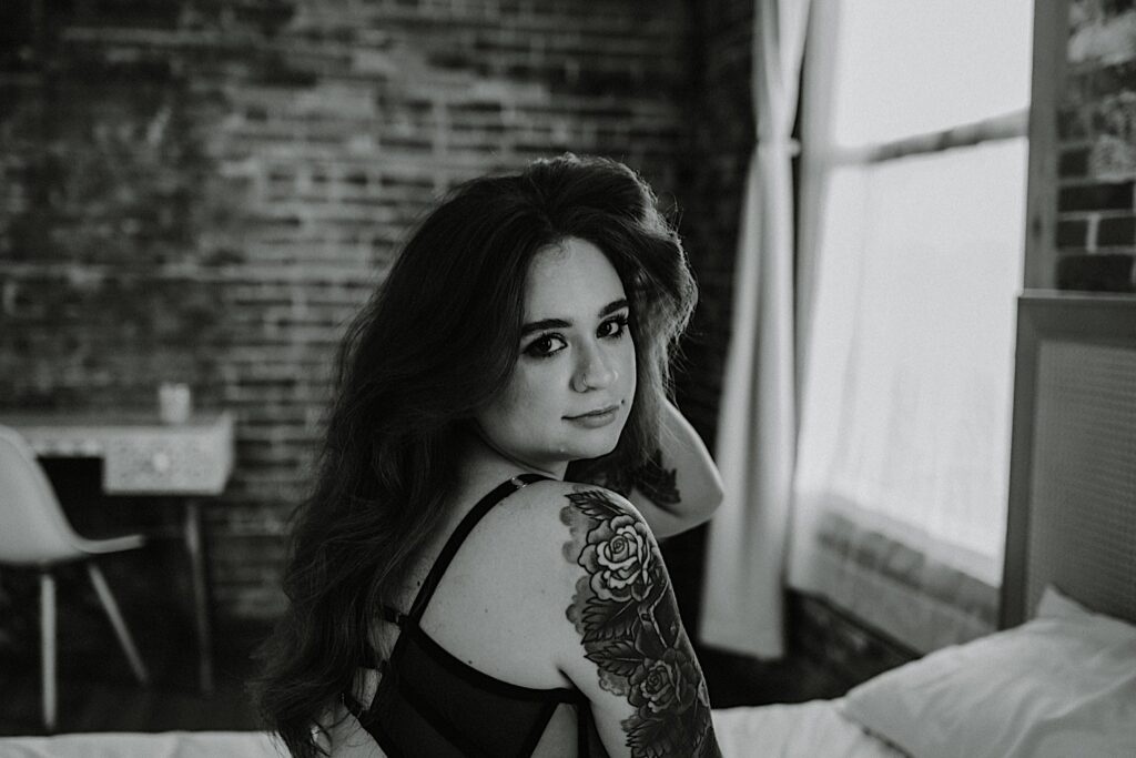 Black and white photo of a tattooed woman in lingerie looking over her shoulder at the camera while having boudoir photos taken of her in Chicago