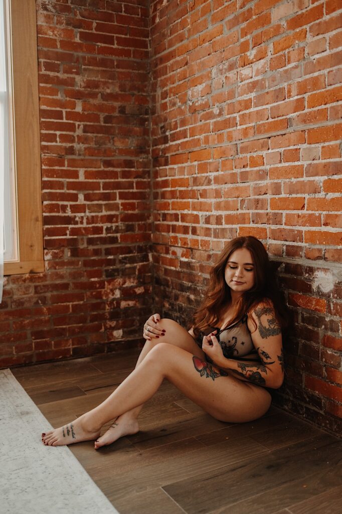 A woman sits on the floor leaning against a brick wall while wearing lingerie during a boudoir photo session