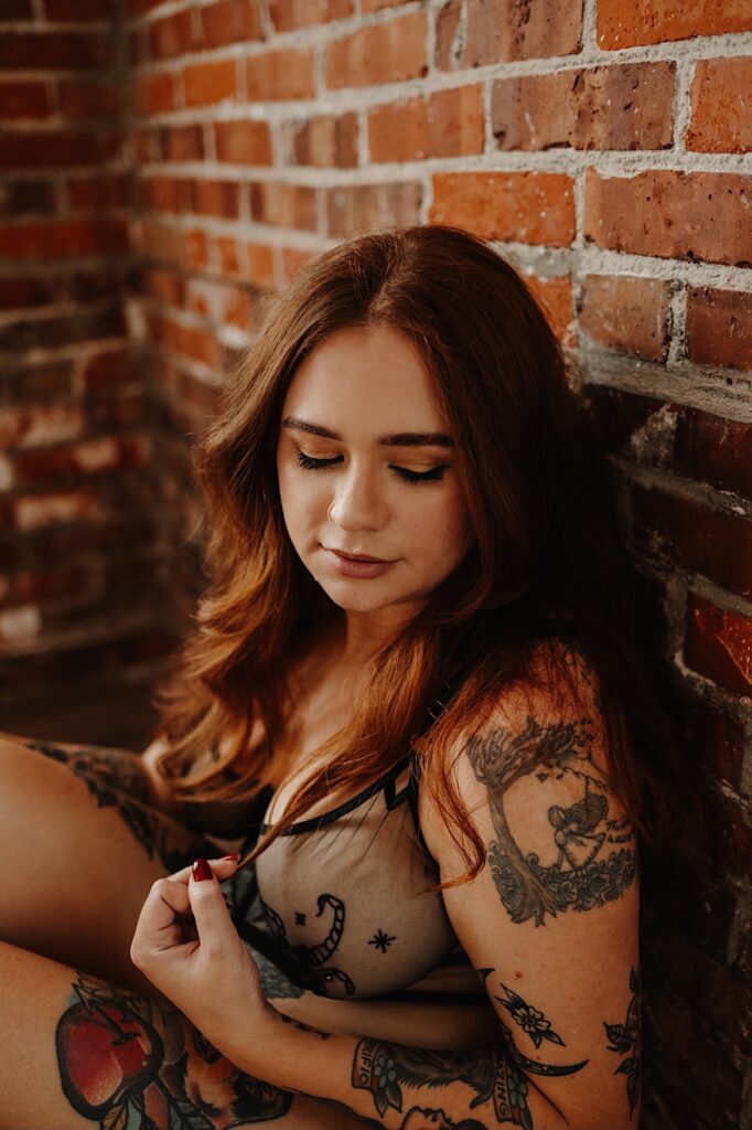 A tattooed woman leans against a brick wall while wearing lingerie and playing with her hair