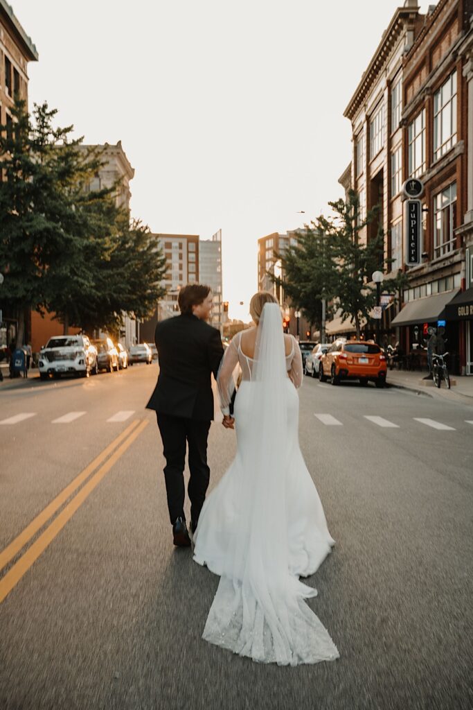 A bride and groom face away from the camera and walk hand in hand down a street in Champaign Illinois at sunset