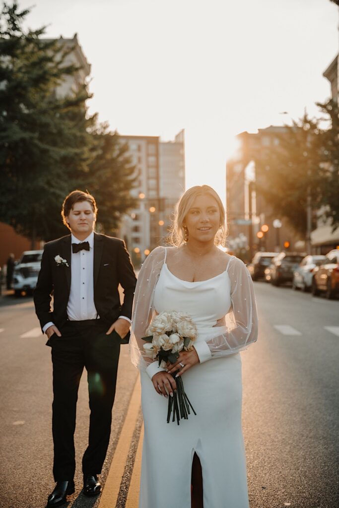 A bride walks towards the camera while the groom watches from behind her as the two stand in a street in Champaign Illinois during sunset