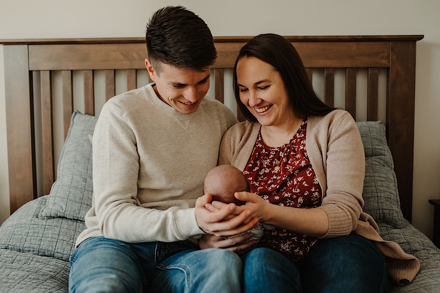 Photo of parents sitting next to one another and smiling as they hold their newborn during their in-home session