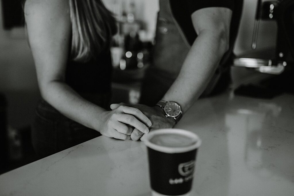 Black and white photo of a couple holding hands facing one another behind a countertop with a coffee cup on it during their engagement session in Bloomington, Illinois