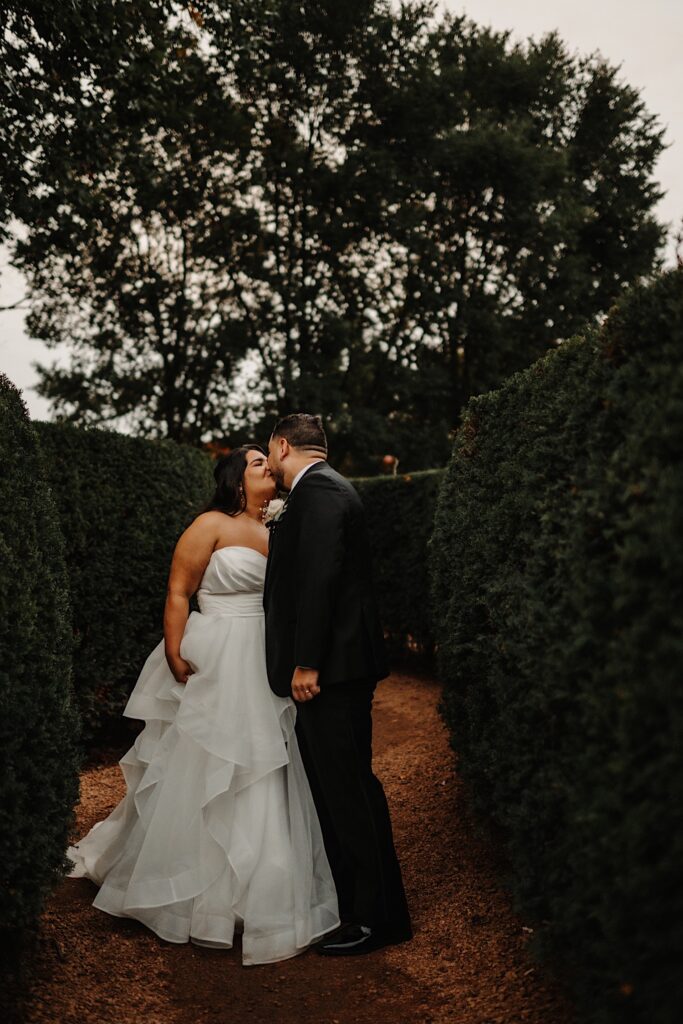 A bride and groom kiss one another while standing outside in a maze of hedges on a cloudy day