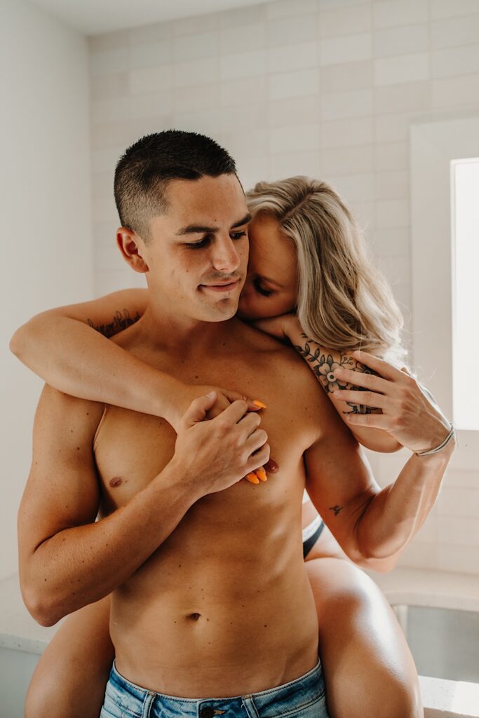 A shirtless man is hugged from behind by a woman sitting on a kitchen countertop
