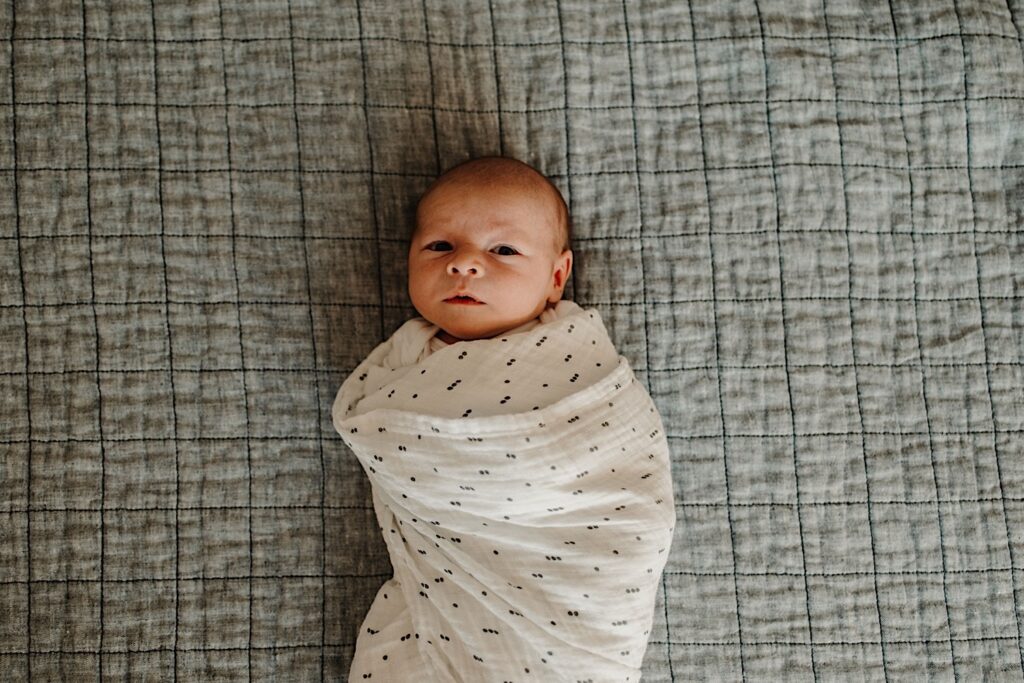 Top down photo of a newborn baby wrapped in a blanket looking up at the camera during an in-home session