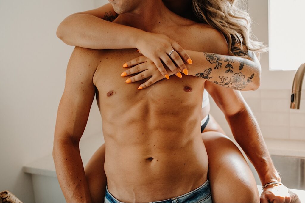 Photo of a shirtless man's torso as he's hugged from behind by a woman sitting on a kitchen countertop in lingerie during a couples boudoir session in the Chicago suburbs