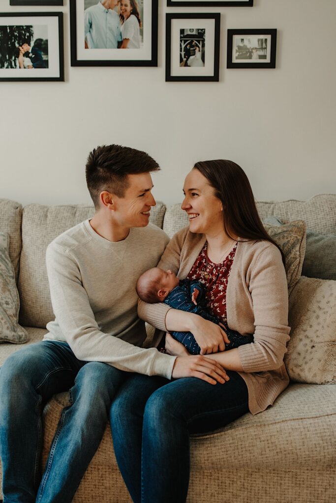Parents smile at one another while sitting on a couch as the mother holds their new baby