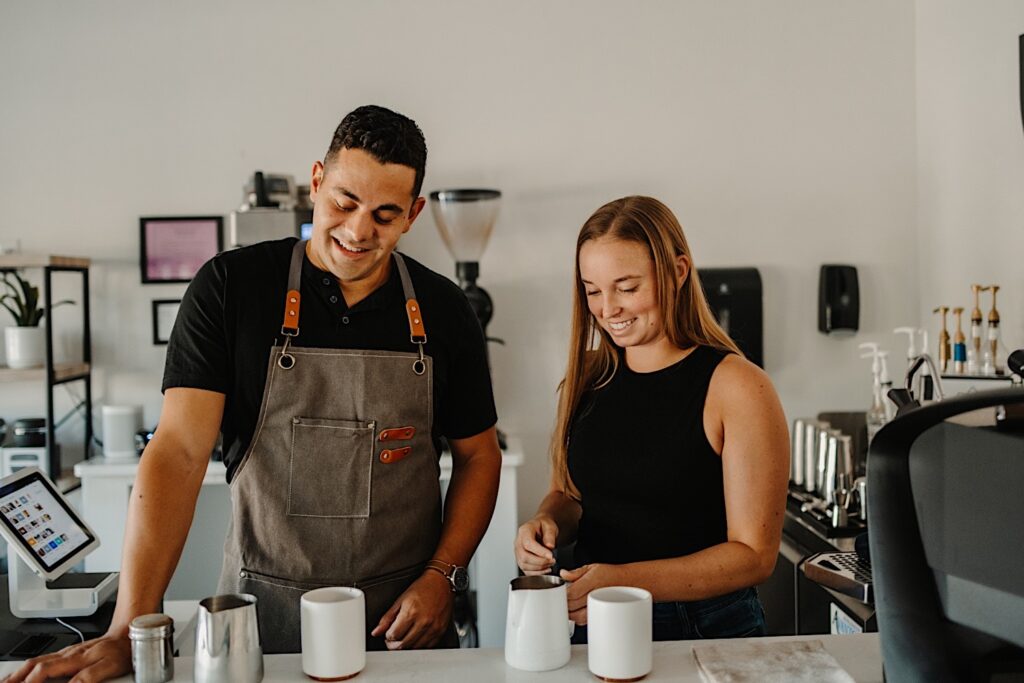 A man and woman stand together in a coffeeshop smiling while making lattes together during their engagement session in Bloomington, Illinois