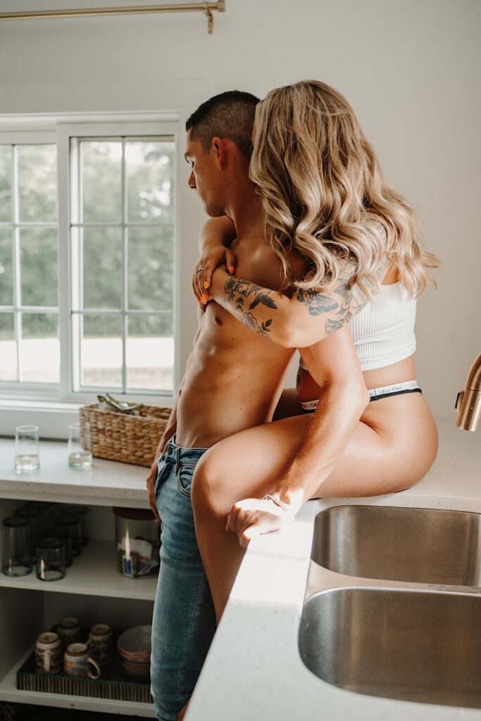 A shirtless man in jeans is hugged from behind by a woman in her underwear sitting on a kitchen countertop