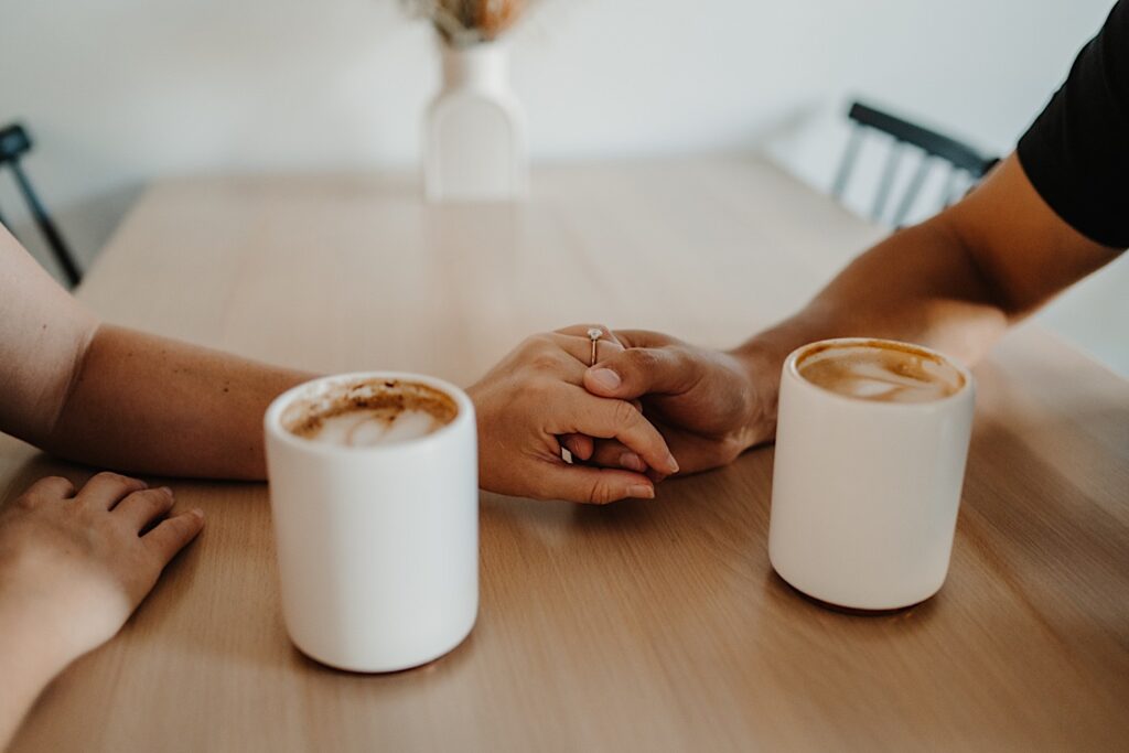 A couple hold hands across a coffee table with lattes resting on the table during their engagement session in Bloomington, Illinois