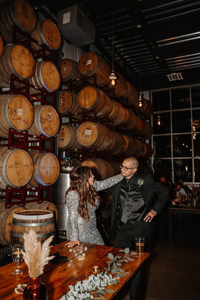 A bride and groom stand next to one another in front of a wall of bourbon barrels as the bride rests her hand on the groom's shoulder