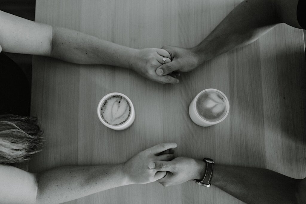 Black and white top down photo of a man and woman sitting across a table and holding hands with one another while two lattes sit on the table between them during their engagement session in Bloomington, Illinois