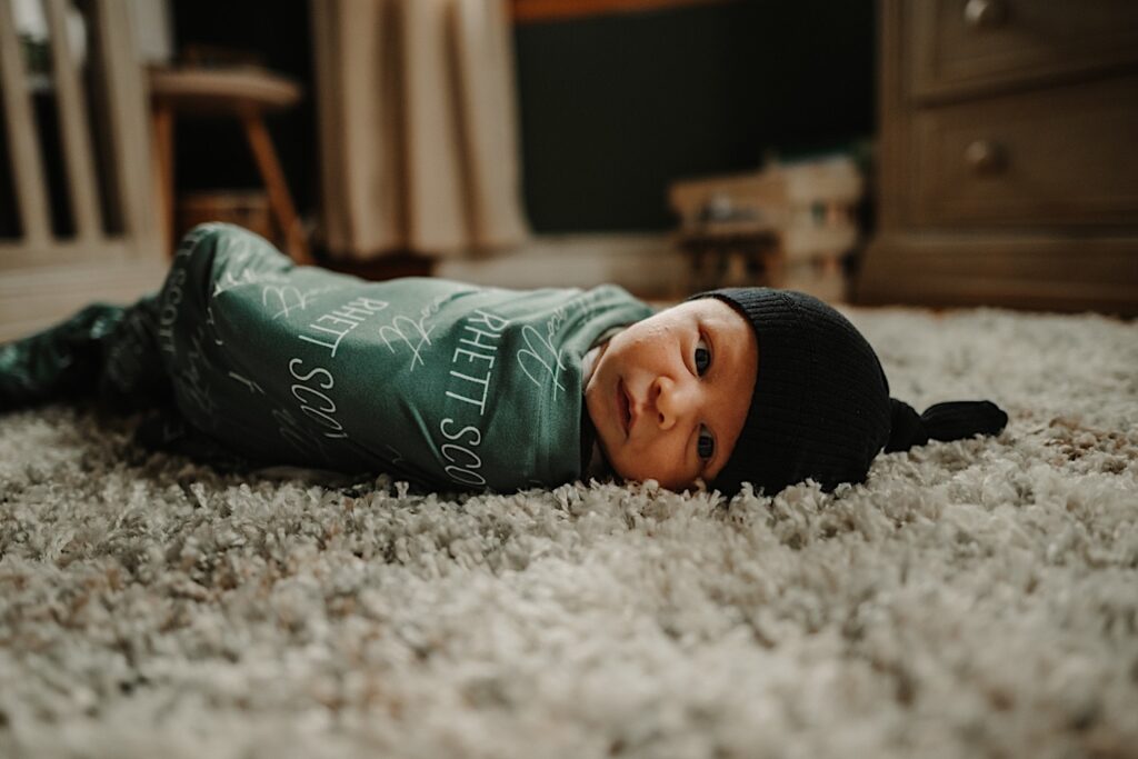A newborn baby wrapped in a blanket looks at the camera while laying on the floor during an in-home photo session