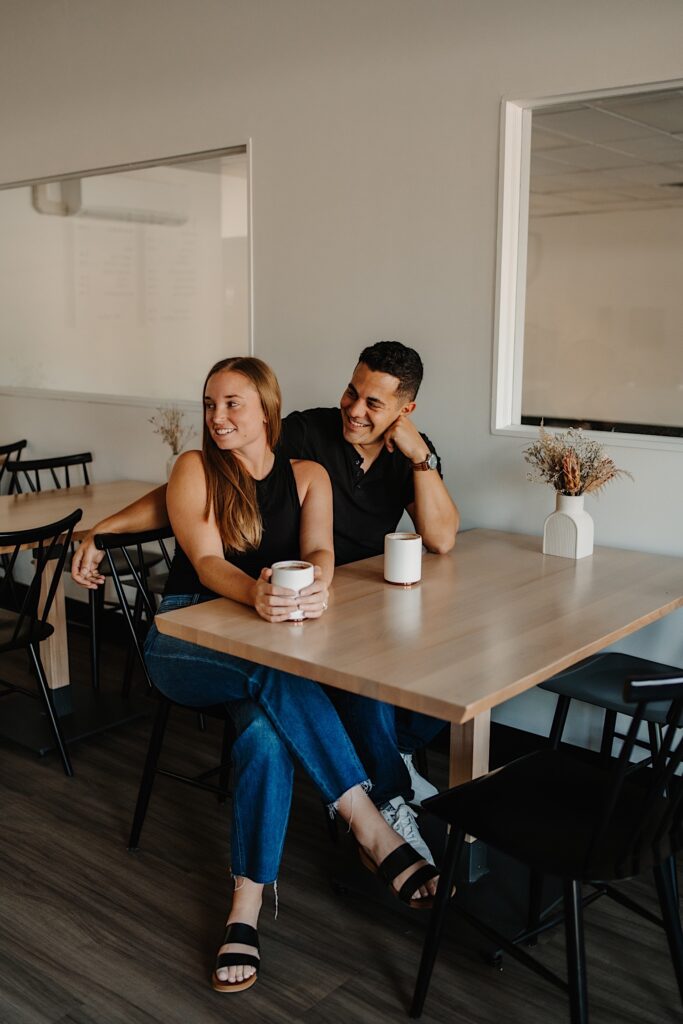 A couple sit next to one another at a table in a coffee shop as they smile to the left