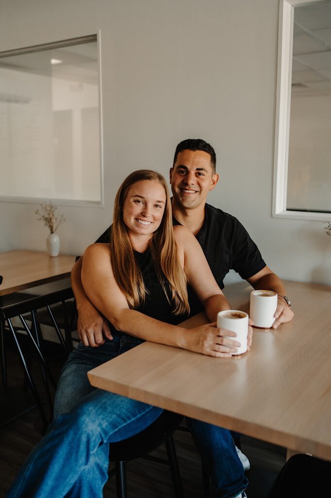 A couple smile at the camera while sitting next to one another at a table in a coffee shop, on the table in front of them are two lattes