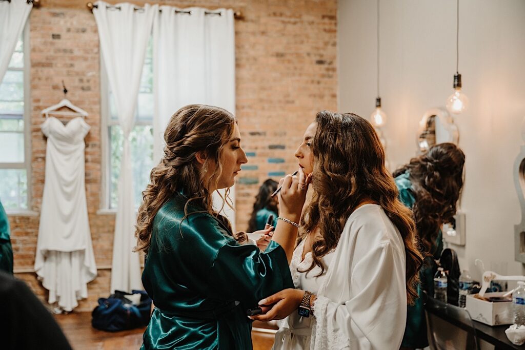 A bride stands in her getting ready space as one of her bridesmaids helps apply her lipstick