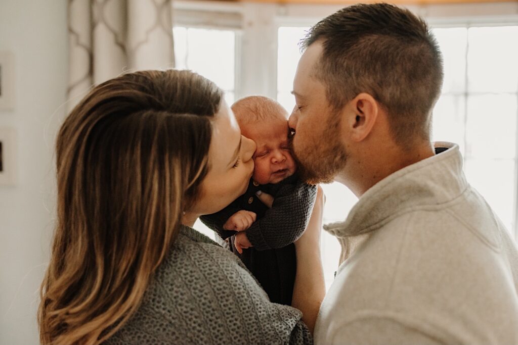 Photo of a mother and father each kissing a cheek of their newborn baby as the mother hold them during their in-home session