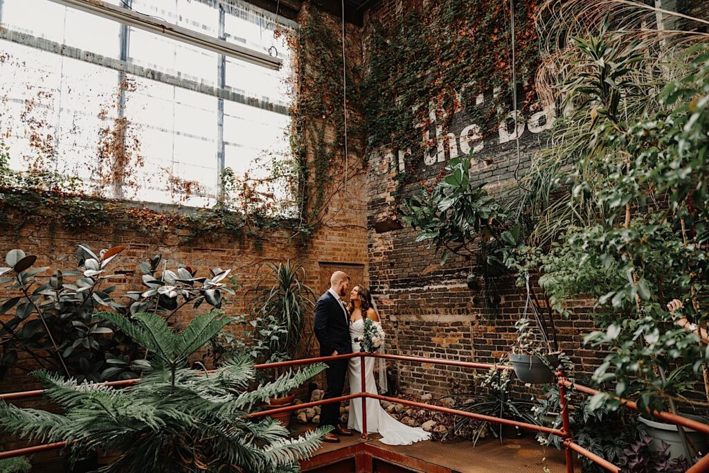 A bride and groom stand side by side and smile at one another while on a metal walkway surrounded by brick walls that are covered in plants and vines