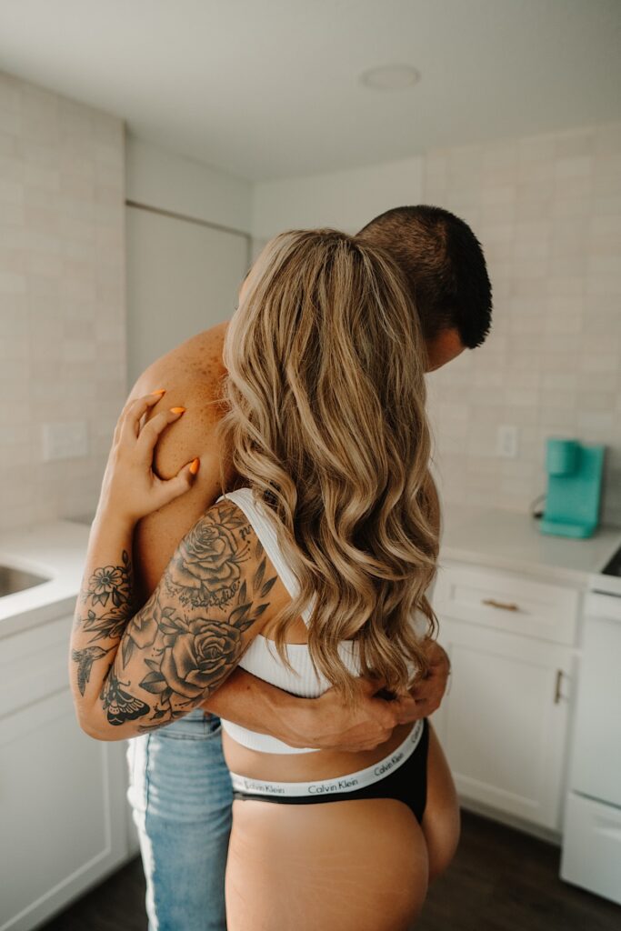 A shirtless man and a woman in her underwear hug one another in the kitchen