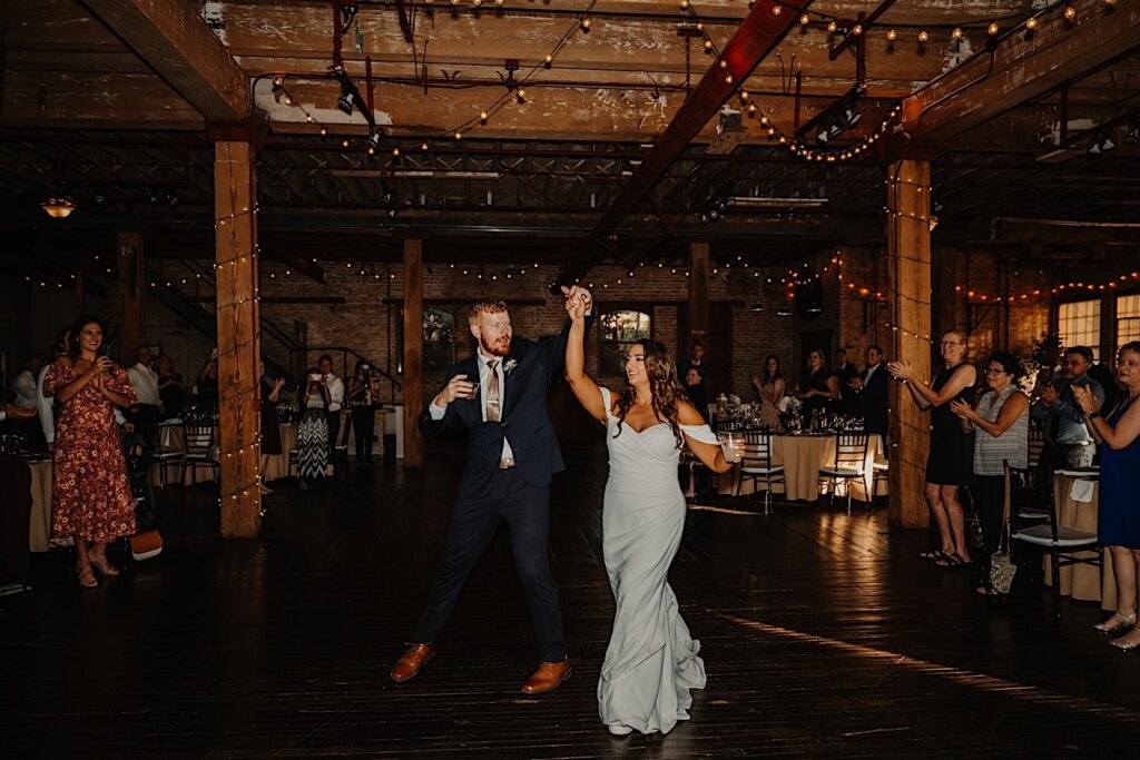 A bride and groom share their first dance together during their indoor wedding reception as guests stand and cheer around them