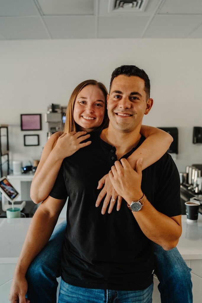 A woman sits on a countertop behind a man and hugs him as they smile at the camera while in a coffee shop