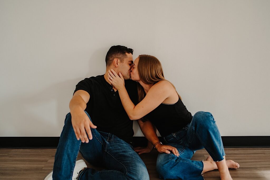 A couple sit next to one another on the floor in a coffee shop and kiss one another during their engagement session in Bloomington, Illinois