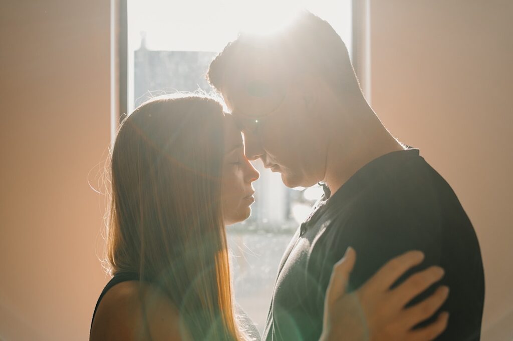 A man and woman stand in front of a window as the sun rises behind them while they touch foreheads together during their engagement session in Bloomington, Illinois
