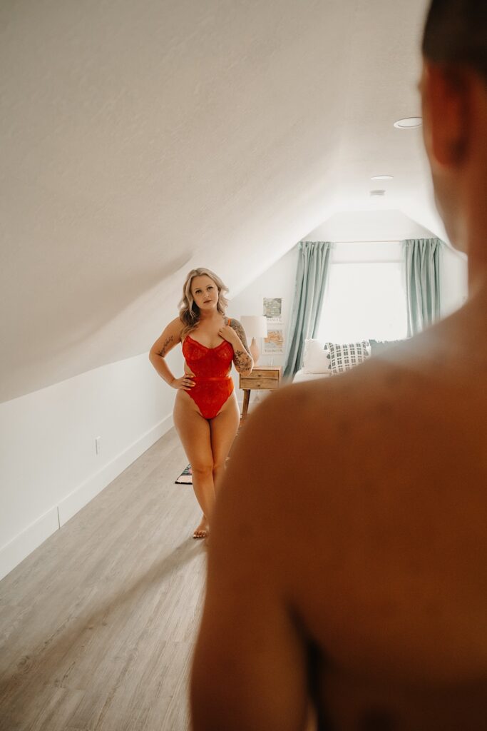 A woman in red lingerie stands in a bedroom, in the foreground of the photo is a man facing her 