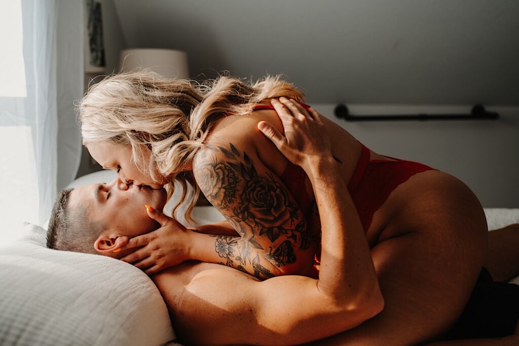 A woman in red lingerie sits atop a man laying in bed and kisses him while he holds her during their couples boudoir session in the Chicago suburbs