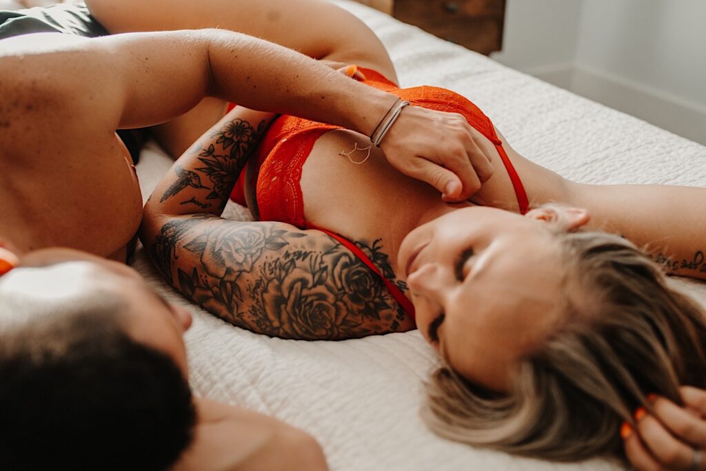 A woman in red lingerie lays on a bed and looks a a man while he touches her chest during their couples boudoir session in the Chicago suburbs