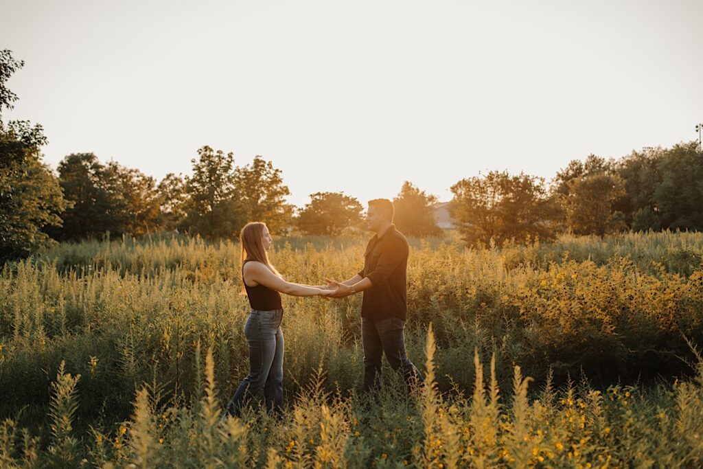 A man and woman hold hands while facing one another in a field as the sun sets behind them during their engagement session in Bloomington, Illinois