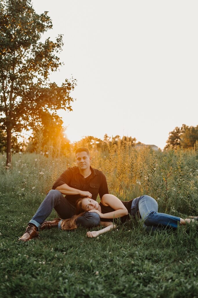 A woman lays on the grass and rests her head on a man's lap who is sitting next to her, the two are in a field with the sun setting behind them