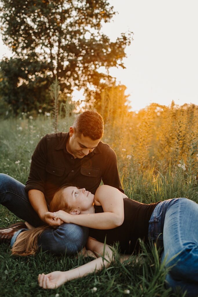 A woman lays on the grass and looks up at a man while her head rests on his lap as he looks down at her while sitting next to her, they're in a field and the sun is setting behind them